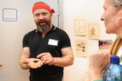 Der Dozent Alex trägt ein rotes Stirnband und zeigt einer Besucherin den Blutegel, den er in seiner Hand hält.
