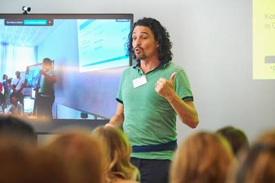 Der Schulleiter Hein Zalorkat hält eine hybride Präsentation über die Ausbildungen an der Heilpraktikerschule Luzern. Er trägt ein grünes T-Shirt. Vor ihm sitzen Teilnehmende, deren Hinterköpfe sichtbar sind – weitere Personen nehmen online teil.