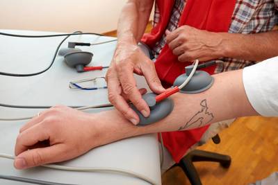Am Methodenbummel kann eine Besucherin unser Elektrotherapiegerät am Unterarm probieren. Das Gerät hat graue Saugnäpfe mit roten und schwarzen Kabeln, die an das Gerät angeschlossen sind.