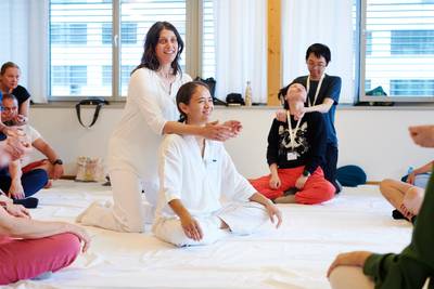 Dozentin Katerina demonstriert eine Shiatsu-Technik an ihrer Kollegin Nina Ninnemann, die im Schneidersitz auf einer Matte sitzt. Gleichzeitig üben die TeilnehmerInnen im hellen Kursraum die gezeigte Technik paarweise miteinander.