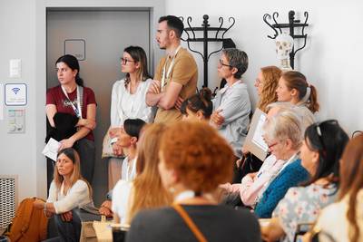 Eine Gruppe von Menschen unterschiedlichen Alters steht und sitzt eng beieinander im Raum 001 und hört aufmerksam dem Kurzreferat zu. Viele tragen Namensschilder um den Hals. Im Hintergrund steht ein menschliches Skelettmodell neben einer Garderobe. 