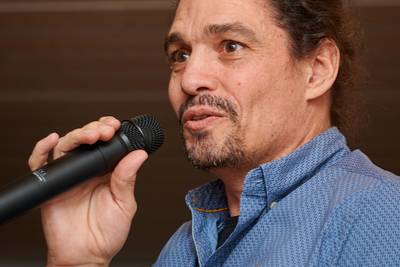 Mann in blauem Hemd hält ein Mikrofon in der rechten Hand und steht vor einer hölzernen Wand mit einer weissen Lampenschirmdecke im Hintergrund. 