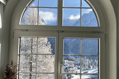 Blick durch ein grosses weisses Fenster mit Rundbogen auf verschneite Berge, Tannen und ein Dorf.