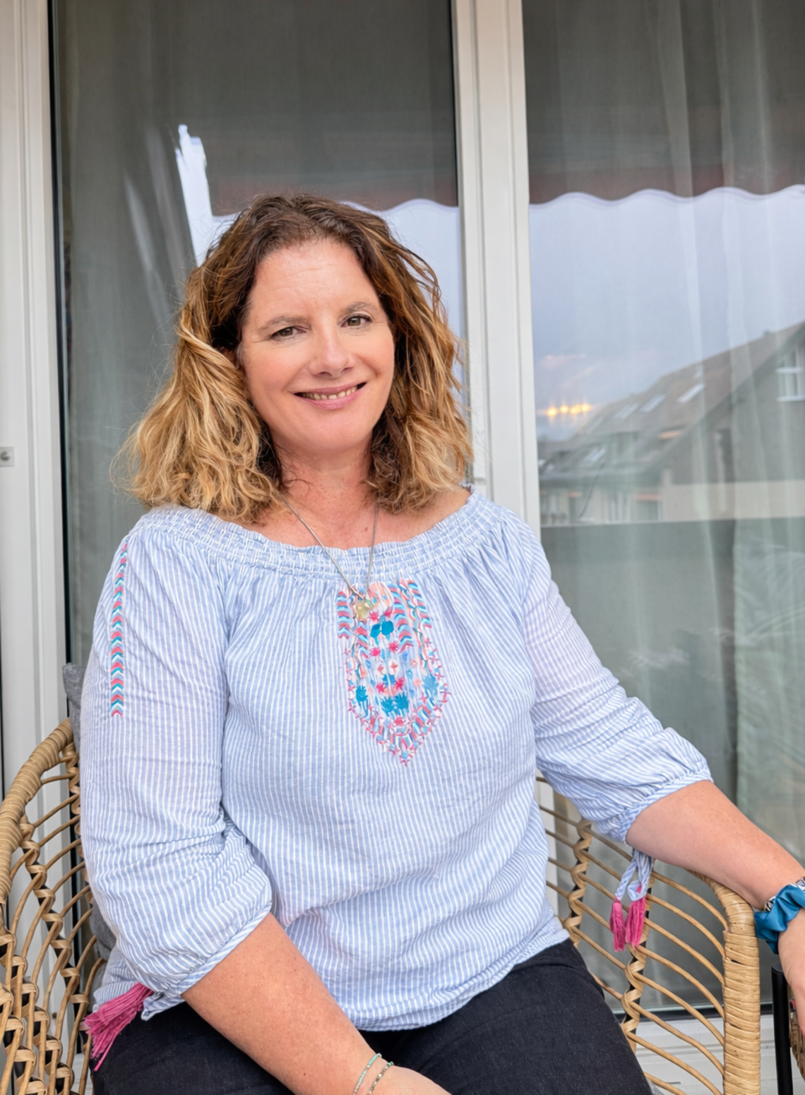 Frau mit schulterlangen, gewellten Haaren sitzt auf einem Korbstuhl vor einem Fenster mit halb geöffnetem Vorhang und trägt eine hellblaue Bluse mit buntem Stickmuster und eine schwarze Hose.