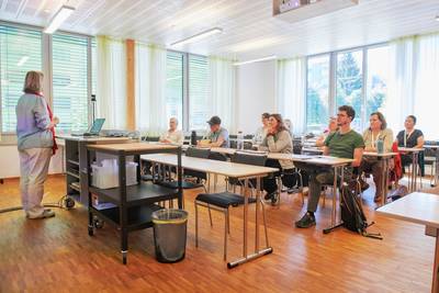 Silvia Schmid steht vor einer kleinen Gruppe, die an Tischen sitzt und ihrem Referat zuhört.