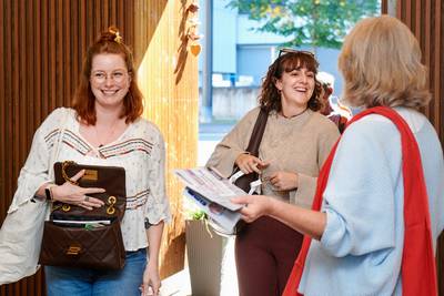Drei Frauen stehen lächelnd am Eingang; eine von ihnen hält Informationsunterlagen in der Hand.