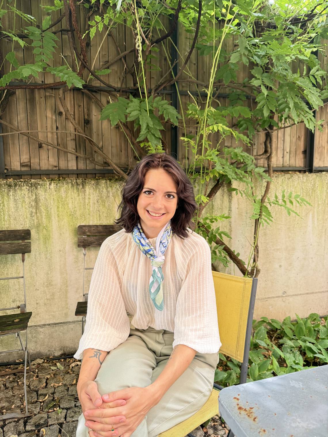 Carol Eltschinger sitzt auf einem gelben Stuhl in einem Garten. Sie trägt eine helle Bluse, ein buntes Halstuch und helle Hose. 