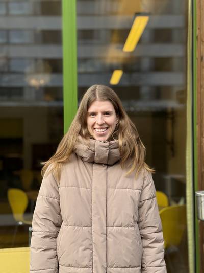 Person mit langen blonden Haaren trägt eine beige Steppjacke und steht vor einem Gebäude mit grünen Fensterrahmen und gelben Stühlen im Hintergrund.