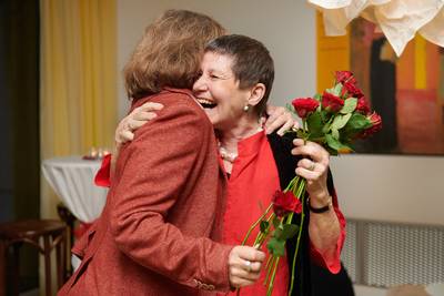 Zwei Frauen umarmen sich, eine hält mehrere rote Rosen in der Hand.