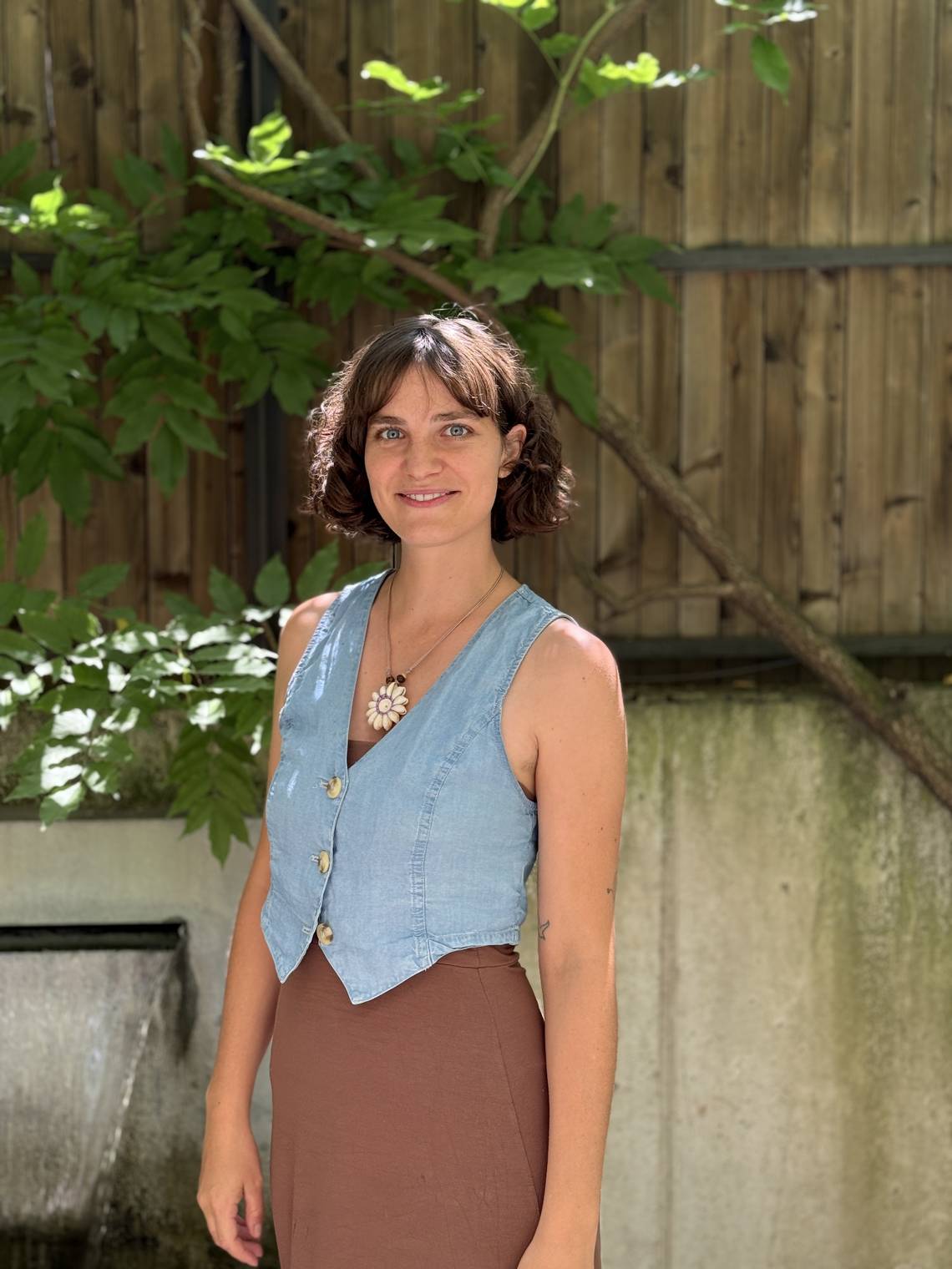 Jana Sieber hat lockiges, braunes Haar und sie steht lächelnd im Sonnenlicht vor einem Holzzaun, umgeben von grünem Blattwerk. Sie trägt ein ärmelloses Jeans-Oberteil, ein braunes Kleid und eine Blütenkette.