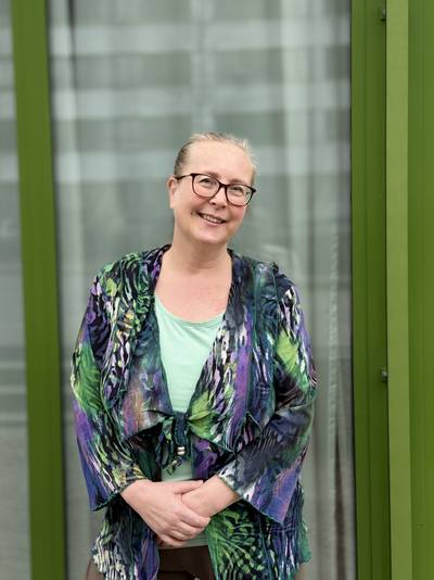 Person mit hellblondem Haar, die vor einem Fenster mit grünen Rahmen steht und eine bunte, gemusterte Jacke über einem hellgrünen Oberteil trägt.