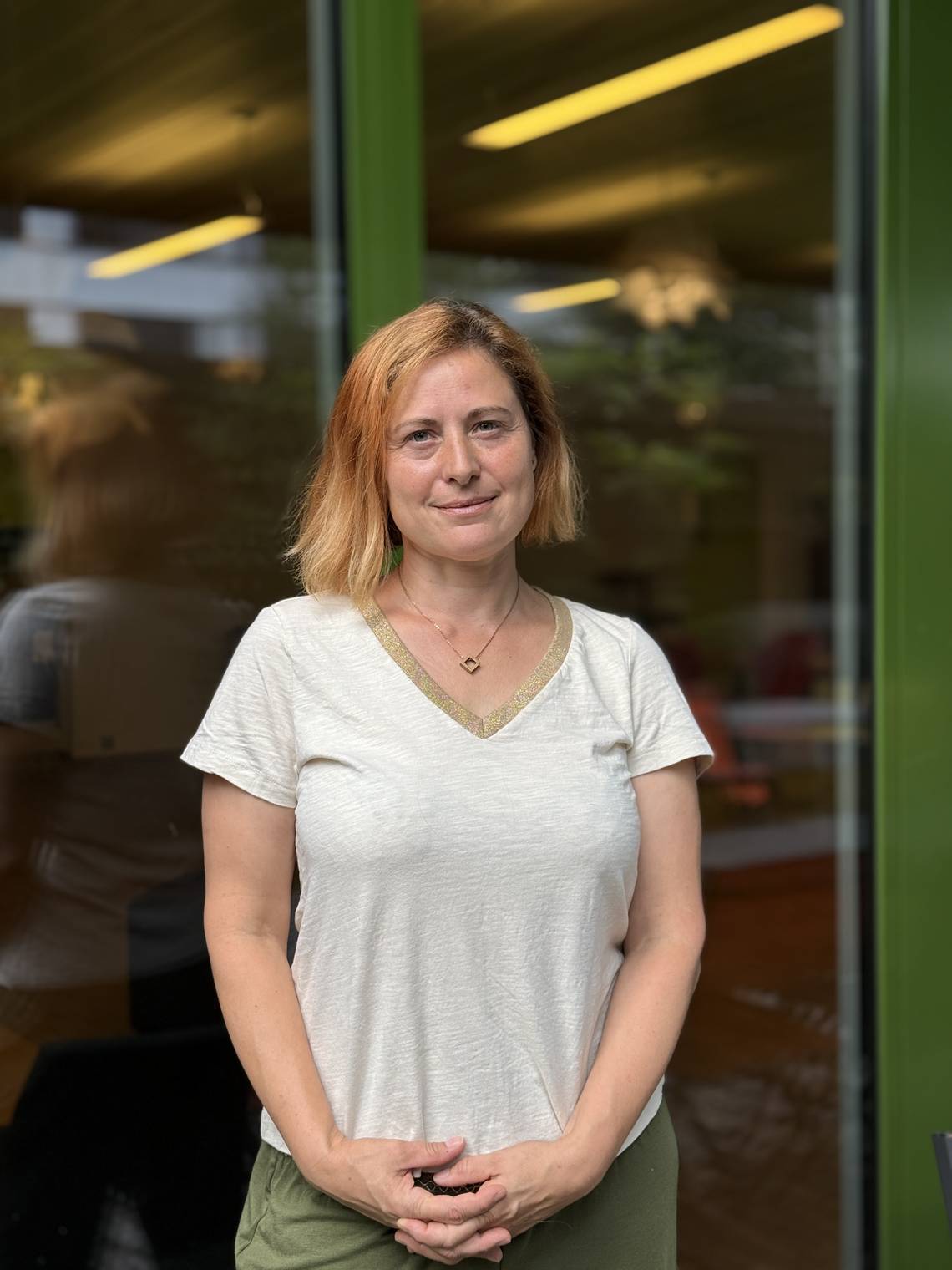 Sonja Keusch steht lächelnd vor der Heilpraktikerschule Luzern vor den Glasfenstern. Sie trägt ein cremefarbenes T-Shirt mit goldfarbenem V-Ausschnitt und eine grüne Hose.