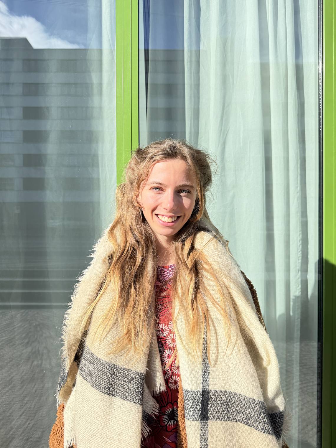 Person mit langen blonden Haaren trägt einen cremefarbenen Schal mit grauen Streifen und ein rotes Oberteil vor einem Fenster mit weissen Gardinen und grünen Rahmen.