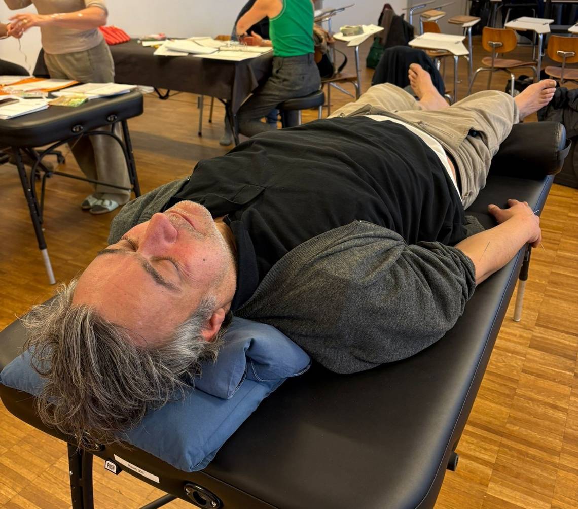 Person liegt auf einer Behandlungsliege in einem Raum mit weiteren Liegen und Personen, die an einem Akupunktur-Unterricht teilnehmen. Foto: Philippe Marty