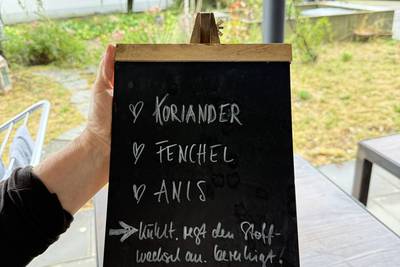 Hand hält eine kleine Kreidetafel mit den handgeschriebenen Zutaten einer Teemischung: «KORIANDER», «FENCHEL» und «ANIS».