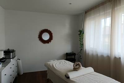 Behandlungsraum mit Massageliege, Handtüchern, Spiegel mit dekorativem Rahmen an der Wand und grossen Fenstern mit Vorhängen.