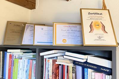 Regal mit zahlreichen Büchern und mehreren gerahmten Zertifikaten auf der Oberseite vor einer weißen Wand und sichtbaren Holzbalken an der Decke.