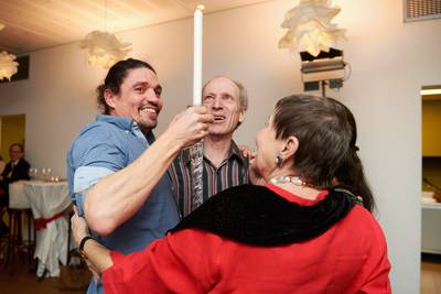 Vier Personen stehen eng beieinander, eine hält eine brennende Kerze hoch, im Hintergrund hängen dekorative Lampen.