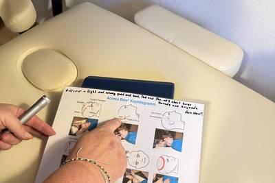 Hand zeigt auf ein Blatt mit farbigen Illustrationen und Texten auf einem hellen Behandlungstisch mit Kopfstütze.