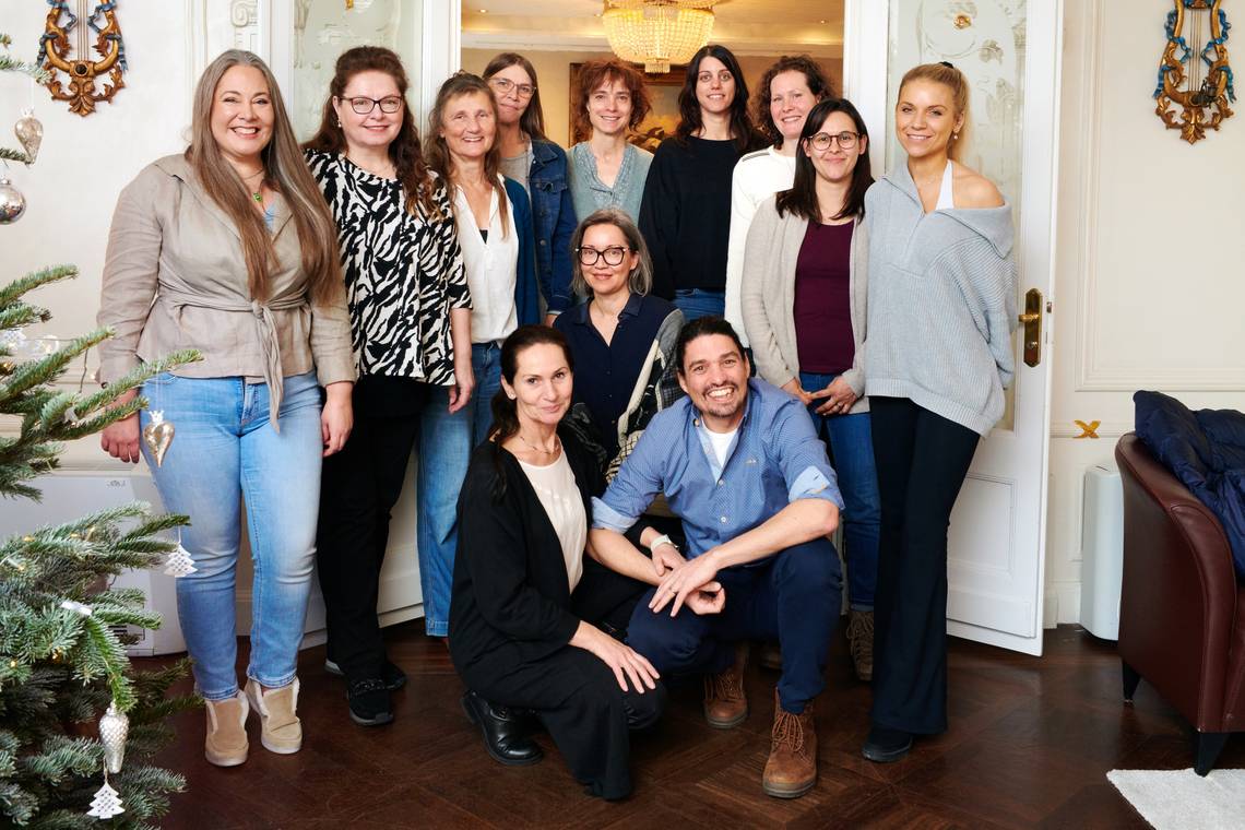 Gruppe von zwölf Personen in einem Raum mit Stuckdecke und Kronleuchter, einige stehen, andere knien. 