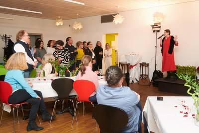 Eine Frau in rotem Kleid spricht auf einer kleinen Bühne vor einem Publikum in einem Raum mit Holzfussboden und weissen Tischen.