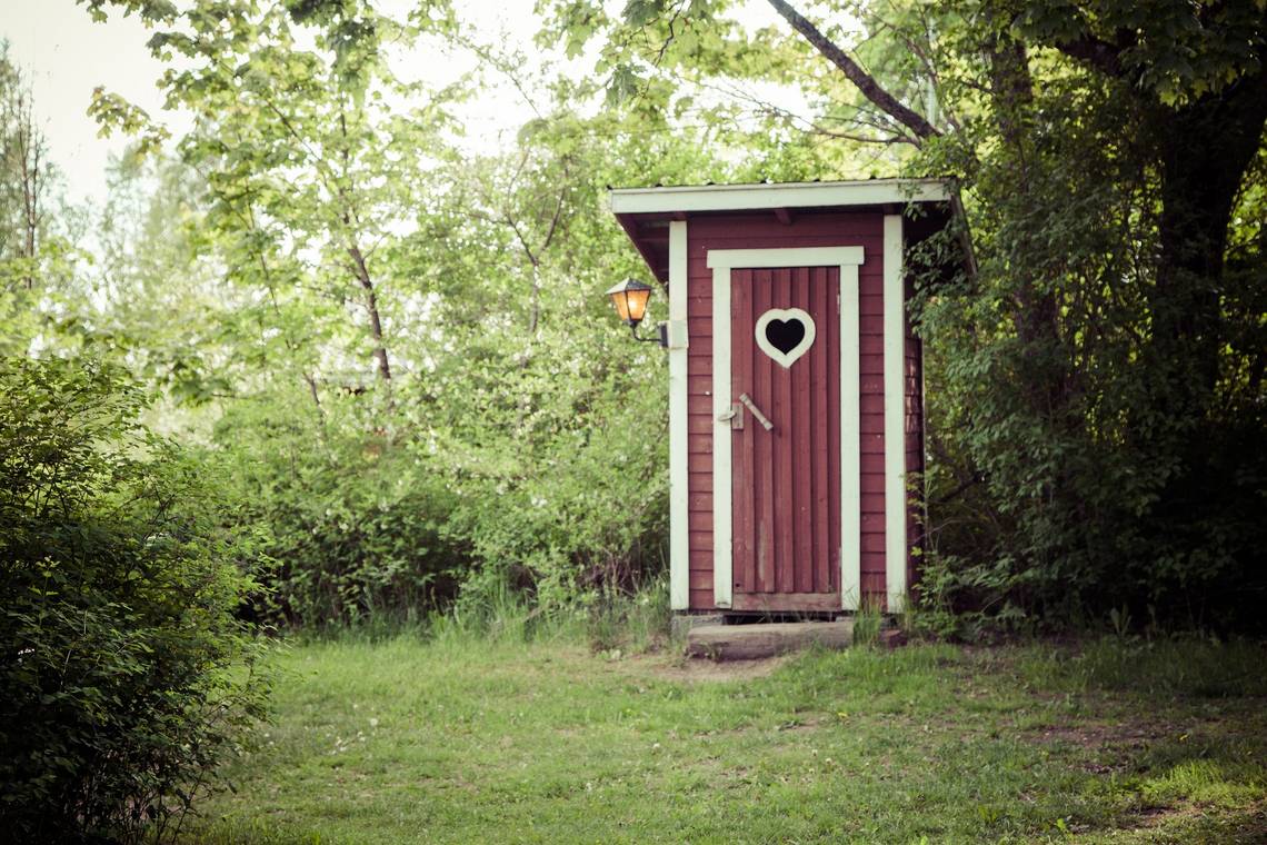 Kleine, rote Garten-Toilette mit weissem Türrahmen und herzförmigem Fenster in einer grünen Waldlichtung