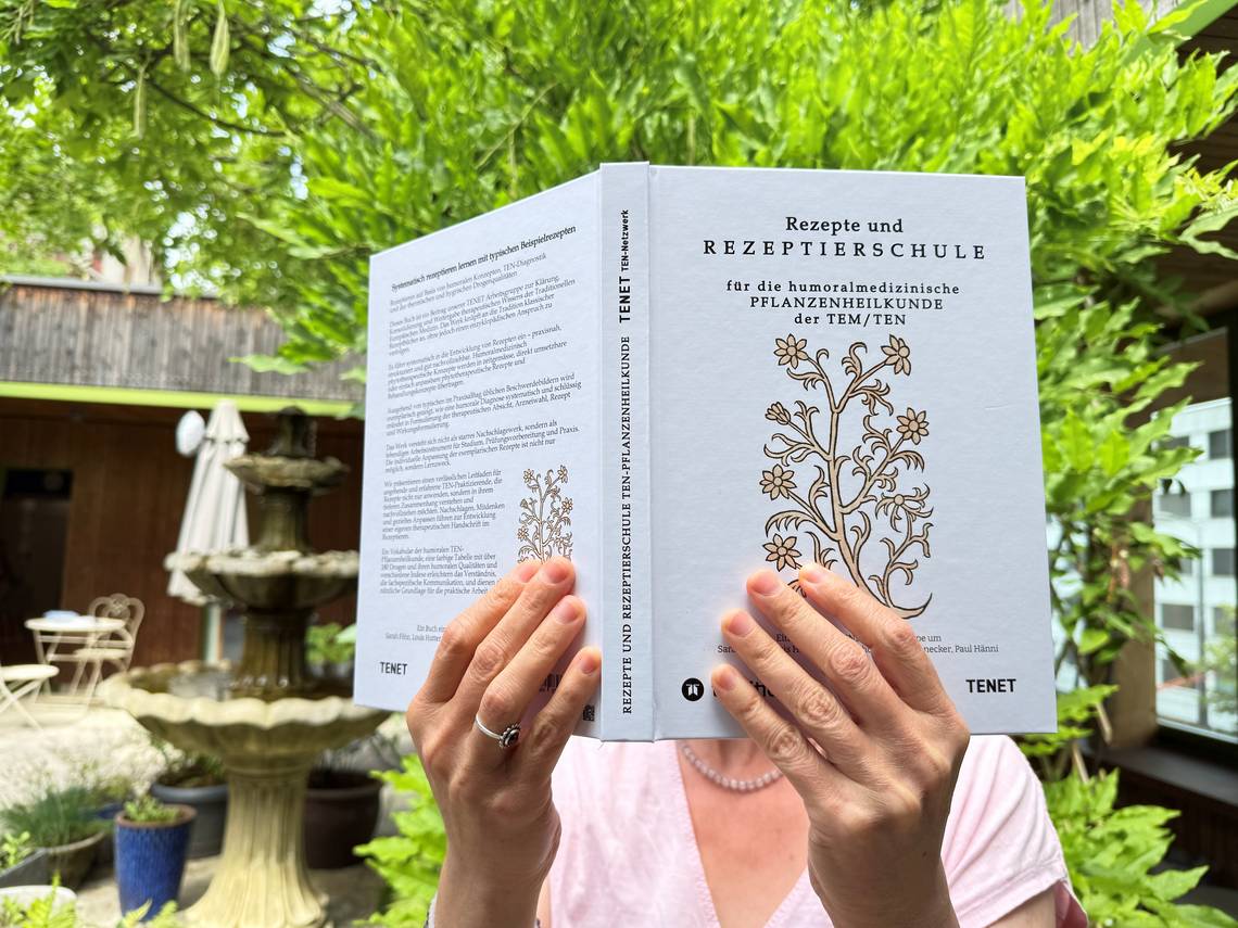 Person hält Buch mit Titel 'Rezepte und Rezeptierschule für die humoralmedizinische Pflanzenheilkunde der TEM/TEN' vor grünem Garten mit Brunnen. Foto: Heilpraktikerschule Luzern