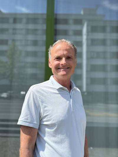 Mann mit weissem Poloshirt steht vor einem Glasfenster mit verschwommenem Gebäude im Hintergrund.
