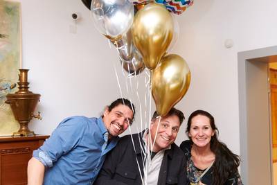 Drei Personen stehen eng beieinander, eine hält einen Strauss aus bunten, goldenen, silbernen und schwarzen Luftballons.