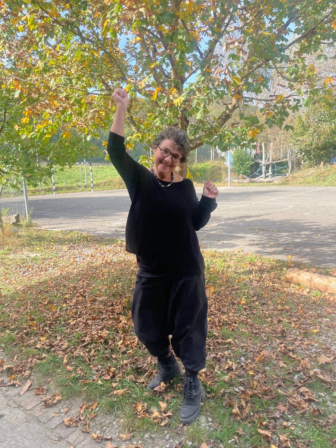 Frau mit Brille und kurzen lockigen Haaren hebt einen Arm freudig unter einem Baum mit herbstlichen Blättern.