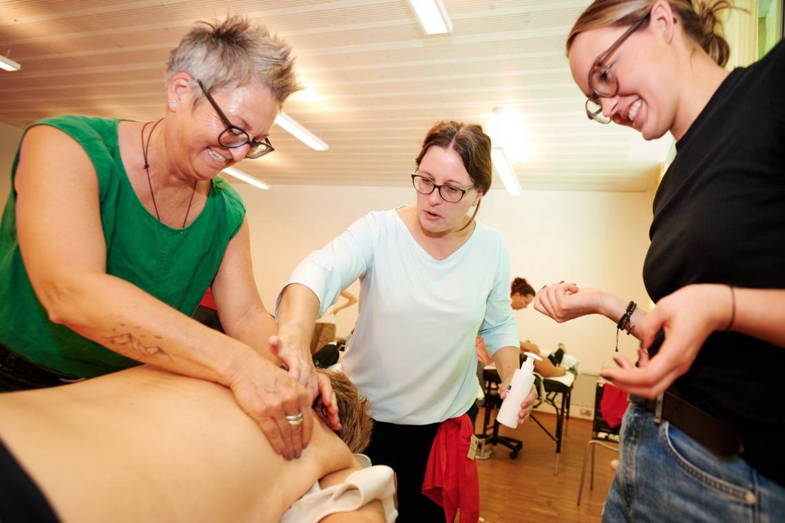 Drei Personen stehen um eine liegende Person, eine massiert deren Rücken in einem hellen Raum; Bild. Maya Jörg