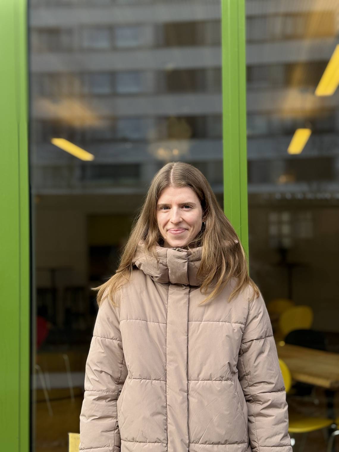 Person mit langen Haaren trägt eine helle Steppjacke und steht vor einer Glasfassade mit grünen Rahmen und gelben Stühlen im Hintergrund.