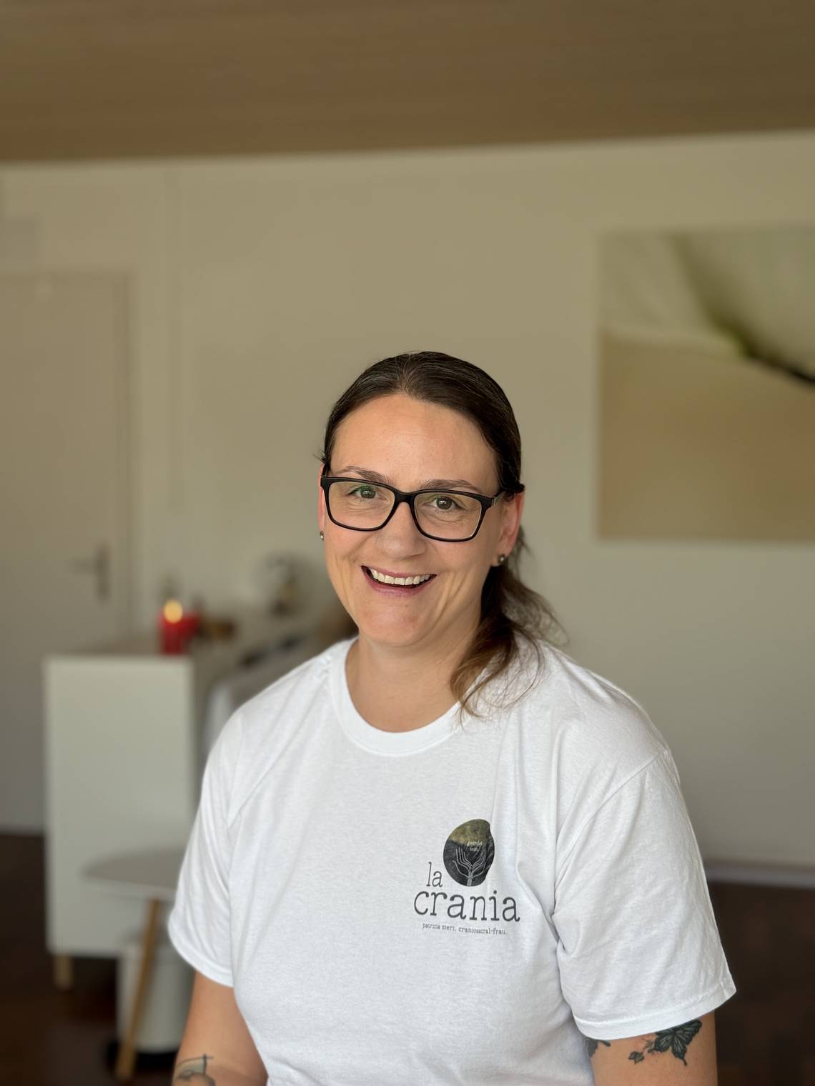 Patricia Zieri sitzt lächelnd in ihrer Praxis. Sie trägt eine weiße T-Shirt mit dem Logo „la crania“. Im Hintergrund ist ein heller, minimalistisch eingerichteter Raum mit einem Bild und einer brennenden Kerze zu sehen.