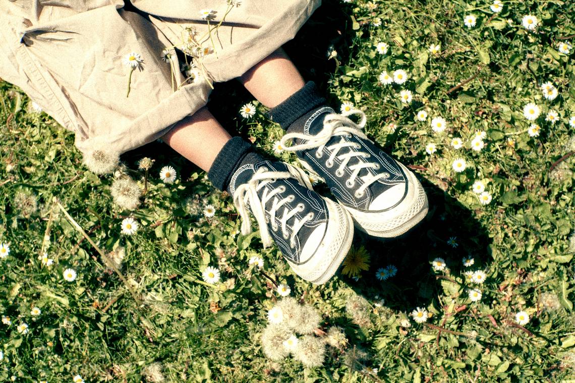 Beine mit schwarzen Sneakers und dunklen Socken liegen auf einer Wiese mit Gänseblümchen und Löwenzahnsamen; Bild: unsplash