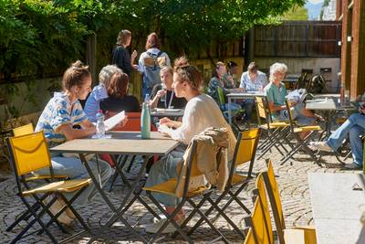 BesucherInnen sitzen draussen bei Sonnenschein an kleinen Tischen und unterhalten sich während einer Pause.