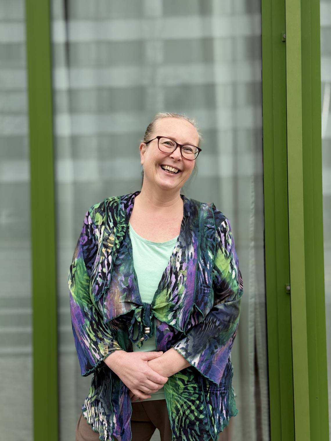 Person mit hellblondem Haar, die vor einem Fenster mit grünen Rahmen steht und eine bunte, gemusterte Jacke über einem hellgrünen Oberteil trägt.