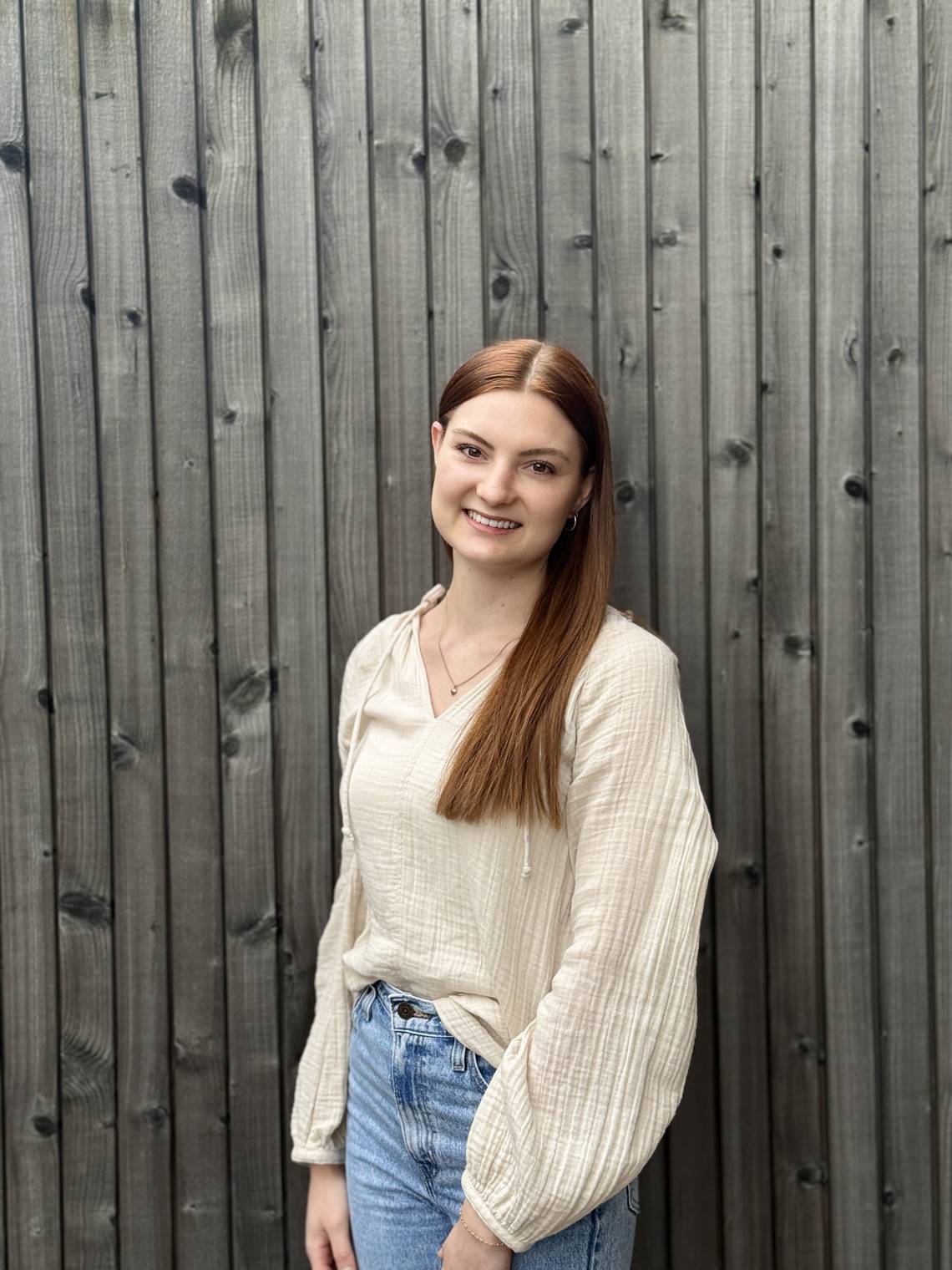 Frau mit langen roten Haaren, weisser Bluse mit weiten Ärmeln und blauer Jeans vor einer vertikal gegliederten Holzwand.