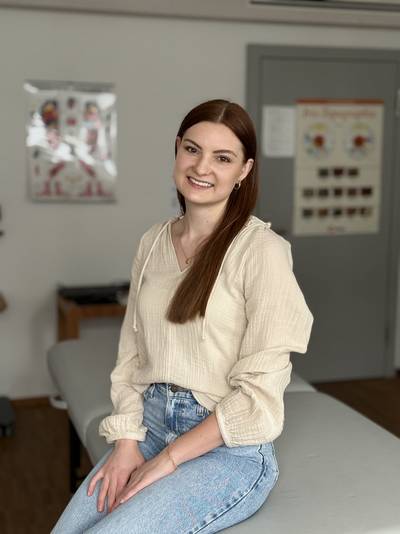 Frau mit langen braunen Haaren, cremefarbener Bluse und blauer Jeans sitzt auf einer grauen Liege in einem Raum mit medizinischen Plakaten an der Wand.