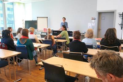 Mehrere Teammitglieder sitzen an Tischen in einem Schulzimmer und hören einer stehenden Person beim Briefing zu.