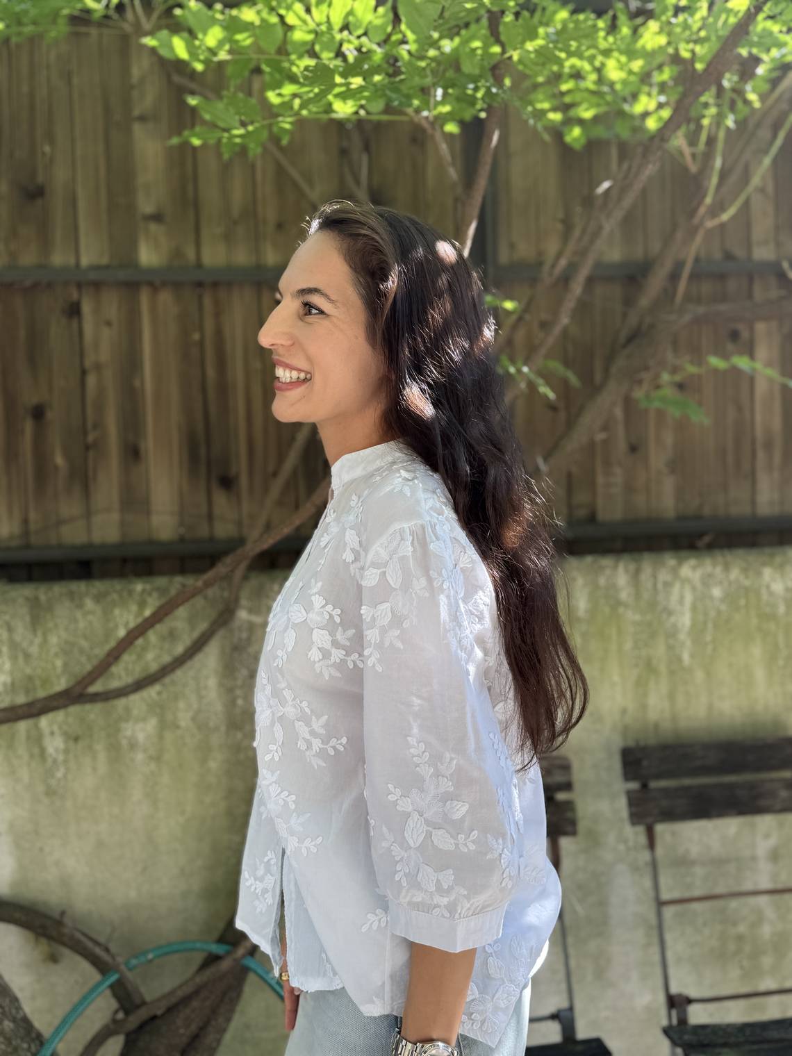 Junge Frau mit langen dunklen Haaren lächelt im Profil, trägt eine weisse bestickte Bluse und steht draussen im Sonnenschein.